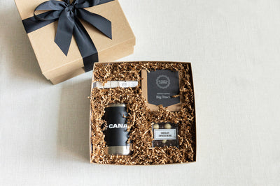 Gift box with shredded paper filler, containing a black mug, coffee package, chocolate espresso beans, and a small box of sugar cubes. The box is tied with a black ribbon.