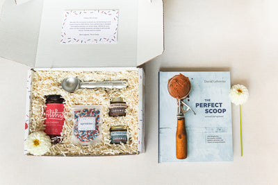 An open gift box contains ice cream toppings, a scoop, and shredded paper filling. Next to it, a book titled The Perfect Scoop by David Lebovitz is on a table with a single flower beside it.