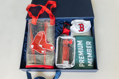 A gift box containing a clear bag with red handles, a small cap with a B logo, a toiletries case, and a green item labeled Premium Member, all featuring a baseball team theme.