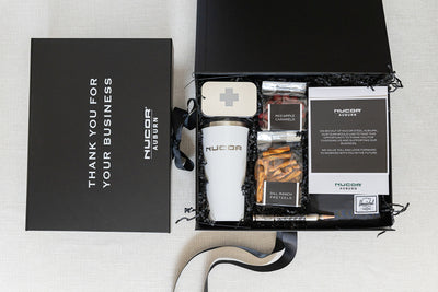 Gift box with Nucor branding showing a tumbler, red apple caramels, pretzels, a pen, and a metal gear coaster, with “Thank You for Your Business” on the lid.