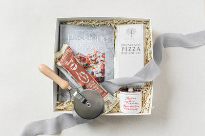 A gift box containing a pizza cookbook, organic pizza flour, pizza cutter, tomato sauce jar, and salami package, all arranged on a light background with a gray ribbon.