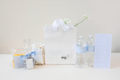 A wedding welcome package with snacks, popcorn, bottled water, and a schedule card in pastel colors is arranged on a table.