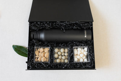 A black gift box containing a black bottle and three clear containers with almonds, round chocolate treats, and powdered candies, all nestled in black crinkle paper. A green leaf is on the left.