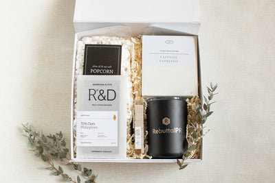 Gift box containing popcorn, chocolate, coffee, and a branded tumbler, surrounded by decorative leaves on a light background.